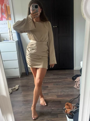 Beige kort klänning med långa ärmar - Trendig beige kortklänning med långa vida ärmar och rynkade detaljer på ena sidan av kjolen. Klänningen har en rund halsringning och markerad midja för en snygg siluett. Perfekt för dig som gillar stilrena och moderna plagg.