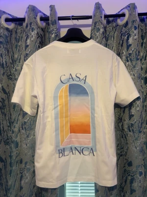 Vit Casa Blanca t-shirt med tryck - Vit t-shirt från Casa Blanca med färgstarkt grafiskt tryck av ett fönster i blått, orange och gult på ryggen samt mindre tryck framtill. Klassisk passform och rund hals. Perfekt för dig som gillar stilrena plagg med unik design.