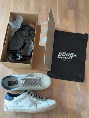 Golden goose superstar - Snygga vita GGDB Superstar sneakers med grå stjärndetalj på sidan, grå skosnören och mörkblå häl. Skorna har en klassisk låg siluett och slitna detaljer för en trendig look. Tillverkade i läder med gummisula. Perfekta för dig som gillar streetstyle.