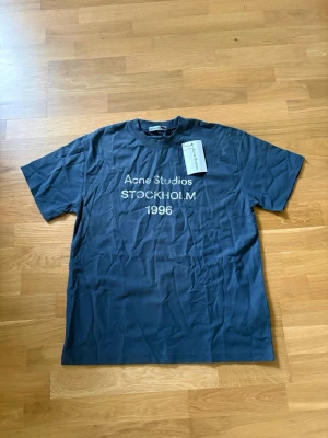 Blå Acne Studios t-shirt med tryck - Cool blå t-shirt från Acne Studios med vitt tryck på bröstet där det står 'Acne Studios STOCKHOLM 1996'. Klassisk rund halsringning och korta ärmar. Perfekt för dig som gillar stilrena och trendiga plagg från svenska designers.