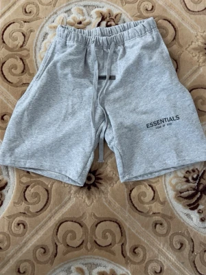 Grå shorts Essentials Fear of God - Säljer ett par grå shorts från Essentials Fear of God. De har snörning i midjan och en relaxed passform. Perfekta för sommaren och tillverkade i mjuk bomull för maximal komfort. Loggan är tryckt på vänster ben.