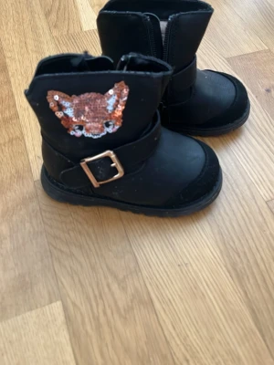 Svarta stövletter med paljettmärke - Säljer ett par svarta stövletter med grov sula och dragkedja på sidan. Dekorerade med ett paljettmärke i form av ett djuransikte och ett snyggt spänne i metall. Tillverkade i syntetmaterial och passar perfekt till en trendig look.