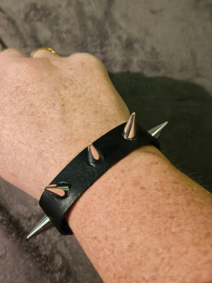 Svart nitar armband med spikes goth Rock Punk  - Coolt svart armband i syntetmaterial med stora silverfärgade spikes längs bandet. Armbandet har tryckknappar för enkel stängning och passar perfekt för dig som gillar en edgy stil.