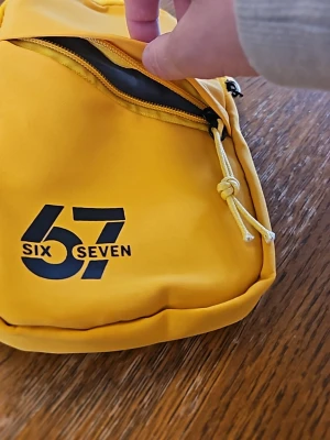 Gul magväska från Six Seven - Snygg gul magväska från Six Seven med svart dragkedja och justerbart band med text. Väskan har ett huvudfack och i huvudfacket finns det ett till litet fack för små saker som pengar nycklar och andra prydlar. Det finns också ett ytterfack.. Materialet är slitstark textil och designen är sportig och trendig.