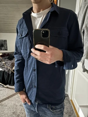 Mörkblå overshirt - Säljer en mörkblå overshirt med två bröstfickor och knappar framtill. Skjortan har lång ärm och en avslappnad passform, perfekt att bära över en t-shirt. Klassisk krage och tillverkad i ett mjukt bomullstyg. Klassisk snygg Grafisk design.