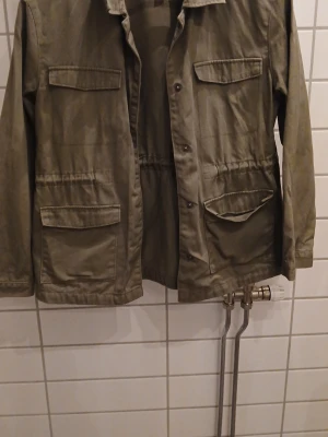 Olivgrön vindjacka med fickor - Snygg olivgrön vindjacka med fyra stora fickor framtill och knappar längs hela framsidan. Jackan har en rak passform och justerbar midja med dragsko. Hör av er angående pris för pris kan diskuteras 