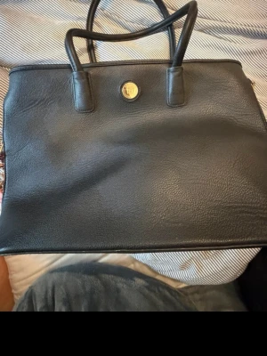 Svart handväska med gulddetalj - Stilren svart handväska i skinn med dubbla handtag och en rund guldfärgad logodetalj framtill. Väskan har en klassisk, fyrkantig form och passar perfekt till alla tillfällen när du vill ha en elegant look.