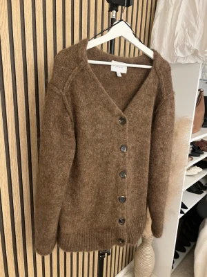 Brun kofta i mohairmix från H&M - Mysig brun kofta i mohairmix från h&m med v-ringning och stora svarta knappar framtill. Koftan är stickad i ett fluffigt material och har långa ärmar samt ribbade muddar vid ärmslut och nederkant. Perfekt för lager på lager och en avslappnad look.