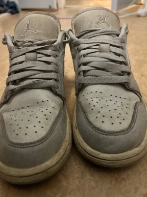 Gråa Air Jordan 1 Low sneakers - Säljer ett par gråa Air Jordan 1 Low sneakers från Nike. Skorna har klassisk låg siluett, perforerad tåbox och ikoniska swoosh-loggan på sidan. Tillverkade i läder med textildetaljer och har broderad Jumpman-logga på plösen.