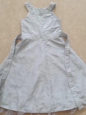 Klänning - Söt silver klänning med broderade detaljer och små paljetter i midjan. Klänningen har breda axelband, knytband i midjan och är tillverkad i ett tunt, lätt tyg. Perfekt för sommarens alla tillfällen. Hällarna är trasiga. Finns fläck.
