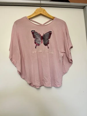Rosa topp med paljettfjäril - Söt rosa topp med lös passform och ett stort paljettbroderi av en fjäril framtill. Under fjärilen står texten 'ONE OF A KIND' . Toppen har korta, vida ärmar och en snygg detalj med korsade band i nacken. Storlek 158/164 eller xs oanvänt 