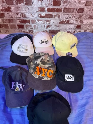 Sju trendiga kepsar i olika stilar - Säljer ett paket med sju kepsar i olika färger och designs. Inkluderar en camo-keps med NYC-tryck, svart keps med WA-logga, gul keps med WA-märke, vit keps med SWEET SKIDS-tryck, svart/vit truckerkeps med ATELIER-tryck, mörkblå keps med H&W-brodyr och en helsvart keps. Perfekt för dig som vill variera din stil.