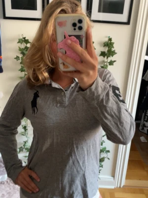 Grå långärmad henley från Ralph Lauren - Klassisk grå långärmad henley från Ralph Lauren med stor svart logga på bröstet och nummerdetalj på ärmen. Skjortan har krage och knappar vid halsen. Tillverkad i mjuk bomull, perfekt för en avslappnad och stilren look.