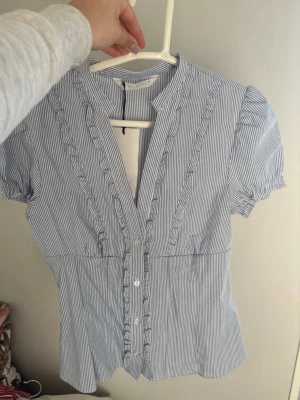 Blårandig blus från Zara - Söt blårandig blus från Zara med korta puffärmar och volangdetaljer längs knäppningen. Blusen har smal krage och knappar framtill. Tillverkad i ett lätt och luftigt tyg, perfekt för en fräsch och stilren look. Aldrig använd, storlek xs. Ny med prislapp
