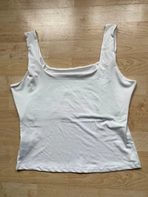 Vit ärmlös linnetopp - Säljer en enkel och stilren vit linnetopp med breda axelband och rak passform. Toppen är ärmlös och har en mjuk, stretchig känsla som passar perfekt till många olika outfits. Perfekt för dig som gillar minimalistisk stil.