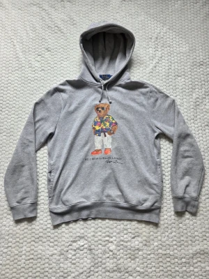 Polo Bear Ralph Lauren Hoodie - Säljer en grå hoodie från Ralph Lauren med den ikoniska Polo Bear-trycket på bröstet. Björnen har på sig en färgglad hawaiiskjorta, vita byxor och röda skor. Hoodien har huva med dragsko och ribbade muddar. Perfekt för en chill och stilren look. Priset kan självklart diskuteras!