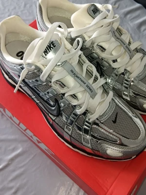Nike silver sneakers p6000 - Nike p6000 sneakers i metallic silver och vitt med svarta detaljer. Skorna har en futuristisk design med mesh och syntetmaterial, tydliga Nike-loggor och vita skosnören. Sitter på en bekväm sula med synlig Air-enhet. Perfekta för dig som gillar statement sneakers. Kommer med boxen. Prisen kan diskuteras. Helt nya inte använda en enda gång. Säljer eftersom skorna efter dom är för småa