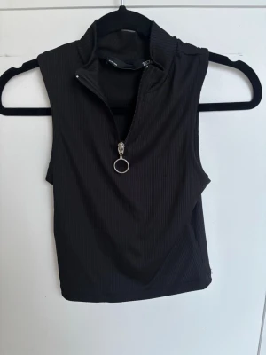 Svart ärmlös crop top med dragkedja - Snygg svart ribbad crop top utan ärmar med hög krage och dragkedja framtill med rund metallring. Perfekt för en trendig och sportig look. Materialet är stretchigt och följsamt.