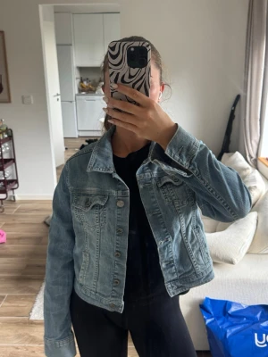 Ljusblå klassisk jeansjacka - Säljer en ljusblå jeansjacka med klassisk passform och snygga detaljer som bröstfickor med knappar och robusta sömmar. Jackan har en cool vintage-look och är perfekt att slänga över en t-shirt. Materialet är jeans och jackan har en rak, kortare modell.