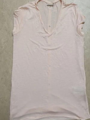 T-shirt - Ljusrosa t-shirt med v-ringning och dekorativa knappar längs hela ryggen. T-shirten har korta ärmar och är tillverkad i ett tunt, mjukt bomullsmaterial. Perfekt för en avslappnad och stilren look.