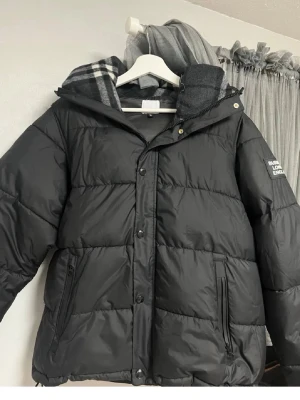 Svart pufferjacka med huva - Säljer en svart pufferjacka från Burberry London England med huva och dragkedja samt knappar framtill. Jackan har två sidofickor med dragkedja och en patch med logga på ärmen. Perfekt för kalla dagar och riktigt snygg streetstil.