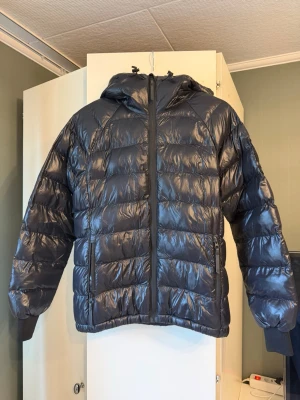 Svart jacka från Peak Performance - Snygg svart dunjacka från Peak Performance med glansig finish, huva och dragkedja framtill. Jackan har breda kanaler, muddar vid ärmslut och en cool detalj på huvan. Perfekt för kalla vinterdagar.