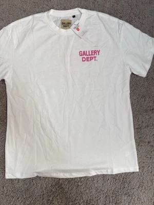 Vit t-shirt från Gallery Dept. - Säljer en clean vit t-shirt från Gallery Dept. med tryckt logga i rosa på bröstet. Klassisk rund hals och korta ärmar. Tillverkad i mjuk bomull som känns skön mot huden. Perfekt för dig som gillar streetwear och enkel stil.