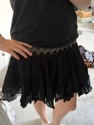 Svart kortkjol med broderad midja - Säljer en svart kortkjol med volang och elastisk midja. Midjan har ett färgglatt broderat zigzagmönster i grönt, rött och vitt som ger kjolen en unik touch. Perfekt för dig som vill ha en skön och snygg kjol till sommaren. Jag har storlek S/M.