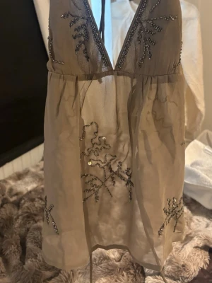Beige glittrig linnetopp med paljetter - Säljer en beige, transparent linnetopp med djup v-ringning och tunna axelband. Toppen har dekorativa paljetter och broderade detaljer framtill och på sidorna🌸