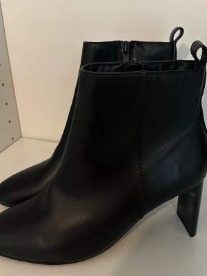 Svarta boots med klack - Snygga svarta boots i slätt skinnimitation med dragkedja på sidan och bred klack. Klassisk och stilren design med rundad tå och ögla bak för enkel påtagning. Perfekta för att lyfta din outfit.