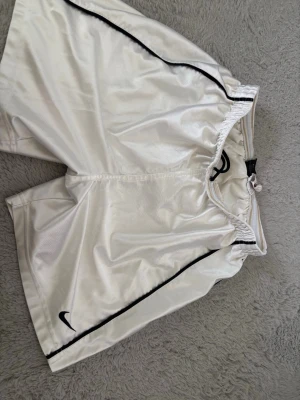 Vita sportiga shorts från Nike - Snygga vita shorts från Nike med svarta detaljer längs sidorna och elastisk midja. Perfekta för träning eller sportiga outfits. Tillverkade i ett glansigt syntetmaterial som ger en modern look. Klassisk Nike-logga på benet. Storlek M