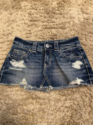 Blå jeanskjol med slitningar - Säljer en kort blå jeanskjol med slitna detaljer och rå kant. Kjolen har fem fickor, bälteshällor och tydliga kontrastsömmar. Perfekt för dig som gillar en avslappnad och trendig stil.