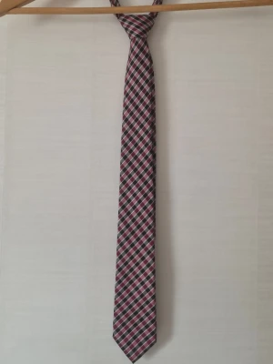 Rutig slips från Bläck - Snygg slips från Bläck med rutigt mönster i vinrött, svart och vitt. Tillverkad i ett glansigt tyg som ger en stilren look. Perfekt accessoar för att lyfta din outfit med lite extra färg och mönster.