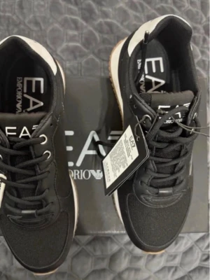 Svarta EA7 sneakers från Armani - Snygga svarta sneakers från EA7 Emporio Armani med vita detaljer och gummisula. Skorna har klassisk låg profil, svarta skosnören och märkets logga på plösen och innersulan. Materialet är en mix av textil och syntet för en modern look.