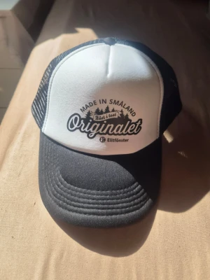 Svartvit truckerkeps Originalet - Säljer en svartvit truckerkeps från Elitfönster med texten 'Originalet' och 'Made in Småland' framtill. Kepsen har böjd skärm, mesh på sidorna och justerbar snapback. Perfekt för dig som gillar klassisk stil och vill sticka ut lite extra.
