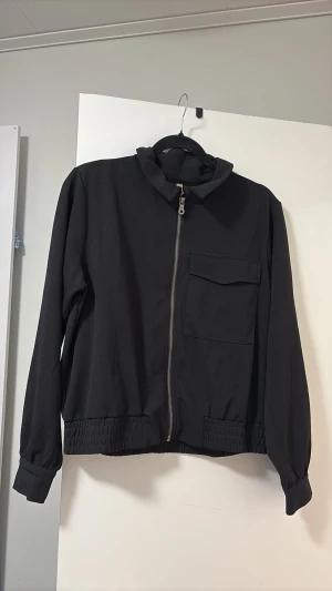 Svart bomberjacka med ficka - Stilren svart bomberjacka med dragkedja framtill och en stor bröstficka med lock. Jackan har ribbade muddar vid ärmslut och midja samt en enkel krage. Perfekt för dig som gillar minimalistisk och clean stil.