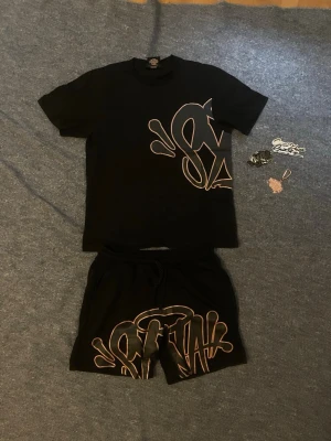 Syna World Set - Svarta shorts från Syna med stort graffiti-inspirerat tryck i beige framtill. Shortsen har elastisk midja med snörning för skön passform. Perfekta för dig som gillar streetwear och vill sticka ut med cool design. Allt inkluderat. 