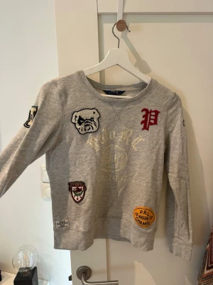 Grå sweatshirt Polo Ralph Lauren - Grå sweatshirt från Polo Ralph Lauren med flera broderade patches, bland annat en bulldogg, ett rött P och olika märkesloggor. 