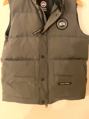 Mörkgrå dunväst från Canada Goose - Snygg mörkgrå dunväst från Canada Goose med klassisk logga på bröstet och två stora fickor framtill. Västen har tryckknappar och dragkedja samt en quiltad design som håller värmen. Perfekt för kalla dagar och enkel att matcha.