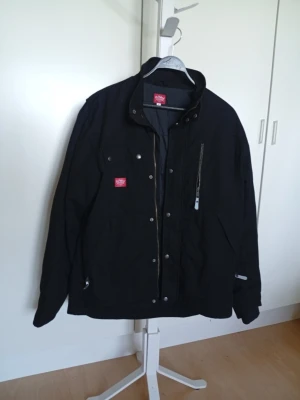 Svart jacka från DePalma - Snygg svart jacka från DePalma med hög krage, dragkedja och tryckknappar framtill. Jackan har flera fickor, inklusive en bröstficka med dragkedja och detaljer i metall. Perfekt för dig som gillar en stilren och funktionell look.