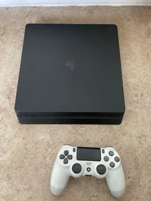 Sony PlayStation 4 Slim med vit kontroll - Säljer en svart PlayStation 4 Slim från Sony tillsammans med en vit DualShock 4-handkontroll. Konsolen har en stilren, fyrkantig design och är tillverkad i matt plast. Perfekt för gaming och streaming av film och serier.