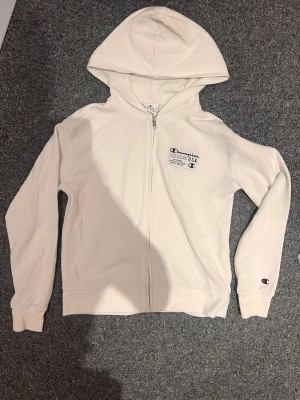 Vit zip hoodie från Champion - Säljer en clean vit hoodie från Champion med dragkedja och huva. Champion-logga på bröstet och ärmen. Tillverkad i mjukt bomullsmaterial, perfekt för en avslappnad och sportig stil. Klassisk passform och ribbade muddar.
