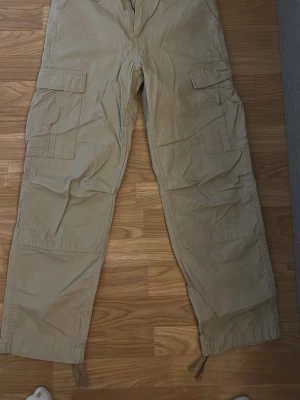 Beige cargobyxor från Carhartt - Säljer ett par beige cargobyxor från Carhartt med flera stora fickor på sidorna och dragsko vid benslut. Byxorna har en avslappnad passform och är tillverkade i slitstarkt bomullstyg. Perfekta för en streetwear-look eller till vardags. Använda en gång. 