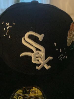 Svart New Era White Sox keps 7 1/4 - Svart keps från New Era med White Sox-logga broderad i vitt framtill. Sidan har broderade blommor i gult, rosa och grönt samt små vita detaljer. Klassisk 59FIFTY-modell med platt skärm och broderade detaljer för en unik look.