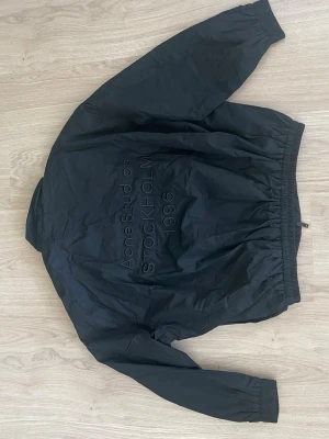 Svart vindjacka Acne Studios - Svart vindjacka från Acne Studios med diskret broderad text 'Acne Studios STOCKHOLM 1996' på ryggen. Jackan har dragkedja framtill, elastiska muddar vid ärmslut och midja samt en minimalistisk design. Perfekt för en clean och stilren look.