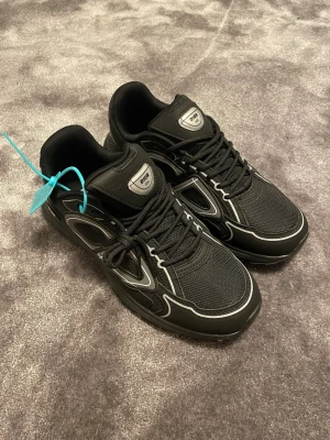 Dior B30 - Snygga svarta sneakers med silverfärgade detaljer och meshpaneler. Skorna har en chunky sula och en cool blå tagg på sidan. Klassisk snörning och sportig design som sticker ut. Perfekta för dig som gillar streetwear och exklusiva märken.