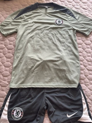 Chelsea FC träningsset från Nike - Chelsea FC träningsset från Nike med matchande t-shirt och shorts. Tröjan är ljusgrön med mörkgrå axelpartier och har Chelsea FC-logga samt Nike Swoosh på bröstet. Shortsen är mörkgrå med vita detaljer och Chelsea-märke. Tillverkad i lätt, ventilerande material.