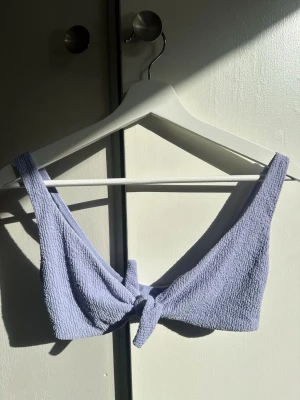 Ljuslila bikiniöverdel med knyt - Trendig ljuslila bikiniöverdel med knyt framtill. Tillverkad i ett strukturerat, frottéliknande material som ger en cool och avslappnad vibe. Breda axelband för extra komfort och stöd. Perfekt för lata dagar på stranden eller poolen.
