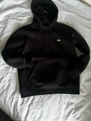 Svart hoodie från Lacoste - Snygg svart hoodie från Lacoste med klassisk krokodillogga på bröstet. Hoodien har en stor magficka framtill. Tillverkad i mjukt material som känns skönt mot huden. Perfekt för en avslappnad och stilren look. Storlek M men är för liten för mig som är 190 liter lång så passar mer som S