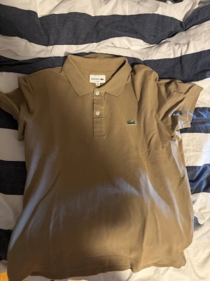 Beige pikétröja från Lacoste - Klassisk beige pikétröja från Lacoste med krage och två knappar framtill. Tröjan har korta ärmar och den ikoniska gröna krokodilloggan på bröstet. Tillverkad i bomull för en skön och luftig känsla.